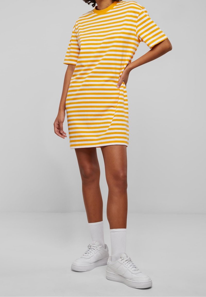 Personne portant une robe à manches courtes rayée jaune et blanche, avec des chaussettes blanches hauteur cheville et des baskets blanches, debout devant un fond gris uni.