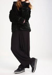 Svart fuskpälsjacka med långa ärmar, lagd över en svart volangtop, kombinerad med vida svarta byxor och svarta skor med vita sulor.