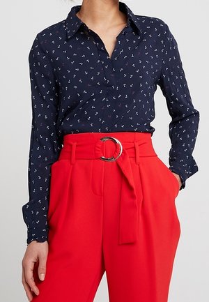 Granatowa koszula na guziki z małymi białymi i czerwonymi wzorami w kotwice, zestawiona z wysokim stanem czerwonymi spodniami z szerokim paskiem z okrągłą srebrną klamrą.