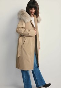 Beżowy płaszcz trencz z futrzanym kapturem, kieszeniami po bokach i paskiem do wiązania. Posiada długi krój i miękką teksturę. Połączony z dzwonami.