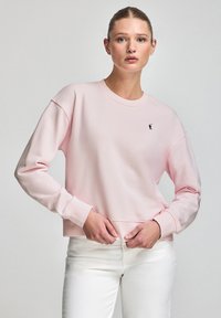 Light pink sweatshirt made of soft fabric, featuring long sleeves, ribbed cuffs, and a small black logo on the chest. Paired with white jeans.