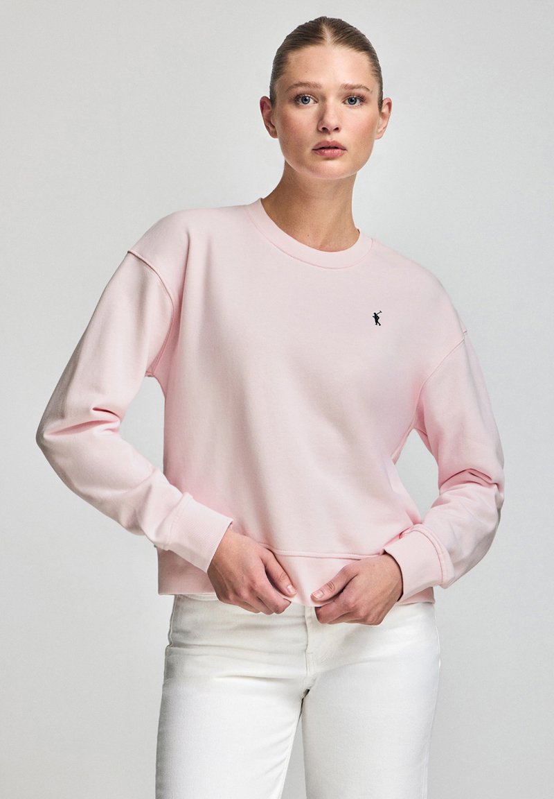 Light pink sweatshirt made of soft fabric, featuring long sleeves, ribbed cuffs, and a small black logo on the chest. Paired with white jeans.