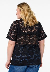 Top in pizzo nero con motivo floreale, maniche corte e un design leggermente trasparente, abbinato a jeans blu.