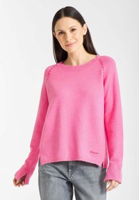 Roze gebreide trui met een ronde hals, zijsplitsingen en geribde manchetten. Heeft subtiele stikdetails op de schouder. Gecombineerd met grijze jeans.