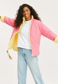 Kapot z prošívaného materiálu s růžovým vnějším povrchem a žlutou podšívkou, se zapínáním na zip a barevnými šňůrkami na táhle zipu.