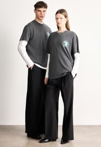 Γκρι υπερμεγέθη μπλουζάκια με λογότυπο, φορεμένα πάνω από λευκές μακριές μπλούζες. Το μοντέλο τα συνδυάζει με μαύρο φαρδύ παντελόνι και κομψά μαύρα παπούτσια.