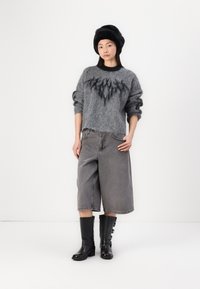 Grijze fuzzy trui met een zwart grafisch patroon, gecombineerd met grijze wijde shorts. Model draagt kniehoogte zwarte laarzen met zijgespen.