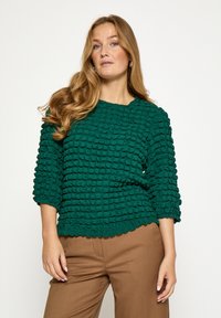 Zelený sveter s textúrou a vzorovaným plyšovým povrchom, s okrúhlym výstrihom a tromi štvrťovými rukávmi, v kombinácii s hnedými nohavicami.