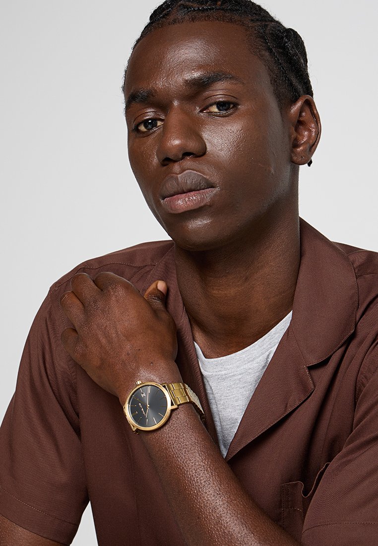 Jeune homme aux cheveux tressés portant une chemise marron et une montre en or avec un cadran noir, reposant sa main sur son épaule.