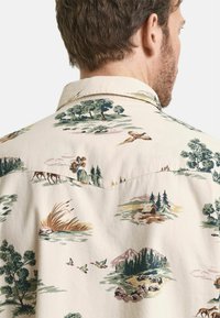 Kurzarmhemd aus leichtem beigen Stoff mit einem detaillierten Landschaftsdruck, der Bäume, Berge und Vögel in verschiedenen Farben zeigt.