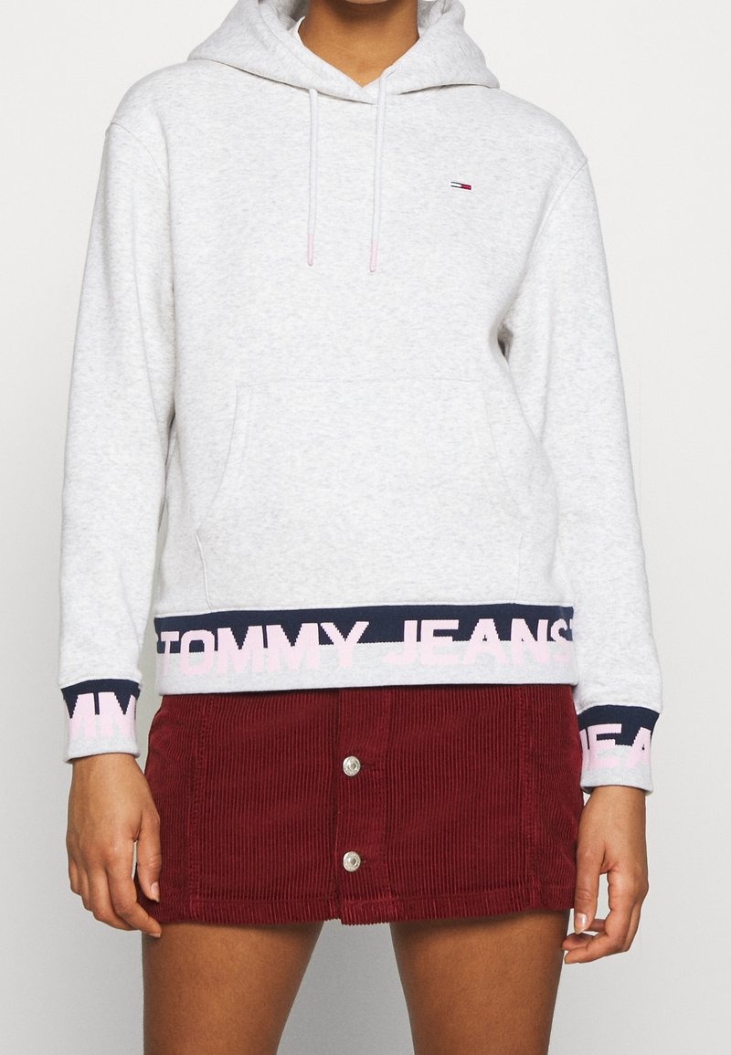 Sudadera gris claro con un bolsillo tipo canguro y puños de canalé azul marino. Presenta una banda rosa con la inscripción "TOMMY JEANS" en el dobladillo. Combinada con una falda de pana roja.
