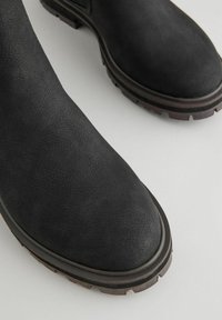 Botas de cuero negro hasta el tobillo con un acabado texturizado, punta redondeada, suela resistente y un patrón de tracción distintivo en un acento marrón oscuro contrastante.