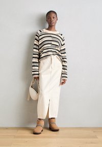 Stribet sort og beige strikket sweater, creme nederdel med en frontslid, brune ankelstøvler med dekorerende bånd, holder en beige håndtaske.