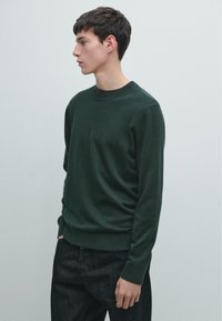 Maglione verde scuro a girocollo realizzato in tessuto a maglia, con maniche lunghe e scollo a coste. Indossato con jeans scuri, larghi e comodi.