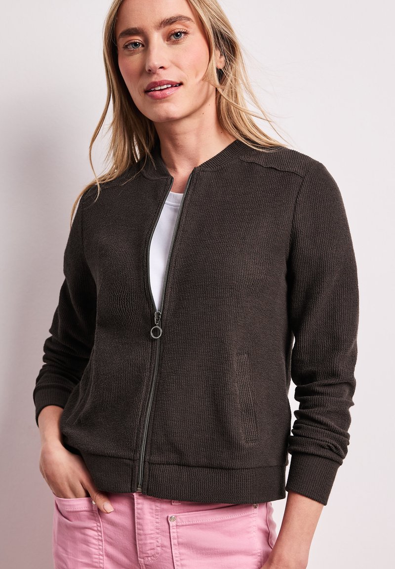 Frau trägt eine dunkelbraune Strickjacke mit Reißverschluss über einem weißen Oberteil und pinken Hosen, stehend mit einer Hand in der Tasche vor einfarbigem Hintergrund.