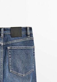Jeans i mellanblå denim med en svart läderlapp på midjebandet. Har en enda bakficka i rektangulär form med kontrasterande sömnad.