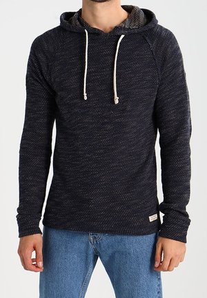 Man wearing a dark textured knit hoodie with white drawstrings and light blue jeans, standing against a plain background.