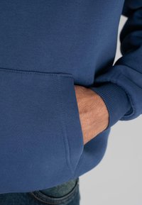Mano insertada en el bolsillo frontal de un sudadera azul con mangas acampanadas, parte de un pantalón de mezclilla visible contra un fondo simple.