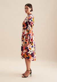 Bunt gemustertes Blumenkleid mit langen Ärmeln, tailliertem Schnitt und ausgestellt Rock. Das Material wirkt glatt, dazu schwarze High Heels.