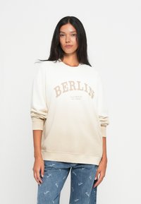 Sweatshirt ombre bege com estampa "BERLIN", gola redonda e punhos canelados. Combinado com jeans azul claro com um padrão de logotipo repetido.