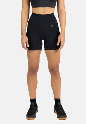 Mujer con pantalones cortos deportivos negros de talle alto, top corto negro, calcetines negros y zapatillas negras con suelas de goma, parada frente a un fondo blanco.