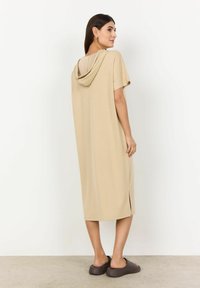 Beige klänning med huva och korta ärmar, sidslitsar och en avslappnad silhuett, kombinerat med bruna slip-on skor på ett strukturerat golv.