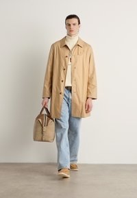 Beige lett jakke med knapper, brukt over en kremfarget turtleneck, kombinert med lyseblå jeans og bærende på en brune veske.