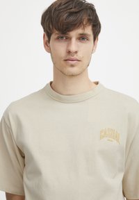 Beige bomull t-skjorte med rund hals, kort ermer, og en gul "CASUAL" brodert logo på venstre bryst. Glatt tekstur.
