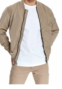 Homme portant une veste bomber beige ouverte sur un t-shirt blanc et un pantalon beige, les mains dans les poches de la veste, torse visible, sans tête montrée.