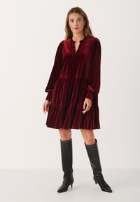 Abito di velluto rosso con orlo arricciato, maniche lunghe e una parte di abbottonatura, completato da stivali neri con tacco al ginocchio.