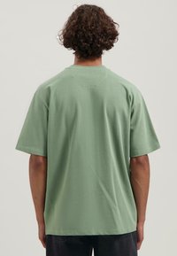 Camiseta verde de manga corta hecha de algodón, con un corte relajado y cuello redondo, que presenta un sutil detalle de costura en la parte posterior.