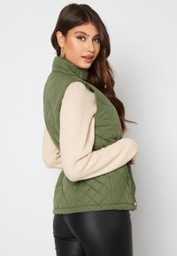 Olivově zelená prošívaná vesta s vysokým límcem, která má boční kapsy a podšitý design, v kombinaci s dlouhým svetrem v jemném béžovém odstínu.