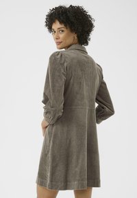 Vestito in corduroy marrone con colletto, maniche lunghe a sbuffo e orlo all'altezza del ginocchio. Caratterizzato da una texture a righe verticali e tasche laterali.