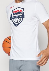 Bílý tričko s krátkým rukávem s logem USA Basketball v červené a námořnické modři, vyrobené z měkkého materiálu, s kulatým výstřihem a standardním střihem.