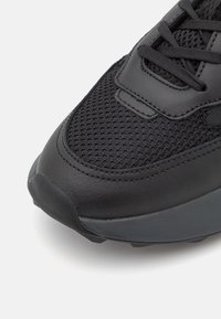 Chaussure de sport noire avec un dessus en mailles, des accents en cuir et une semelle épaisse en caoutchouc avec une adhérence texturée. Détails de conception minimalistes.