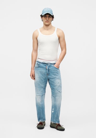 Νέος άνδρας που φοράει λευκό φανελάκι, ανοιχτό μπλε ταλαιπωρημένο τζιν, πράσινα αθλητικά παπούτσια και ανοιχτό μπλε καπέλο με την επιγραφή "ROOT".