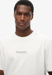 Camiseta de algodón blanca con una textura suave, con cuello redondo y el logo "Superdry." en gris en el centro. Mangas cortas y ajuste relajado.