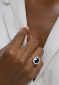 Sapphire ring with a silver band, oval center stone surrounded by small diamonds, set against a textured skin backdrop and white fabric.