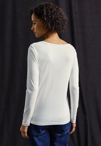 Chemise blanche à manches longues avec un col large, texture lisse et design ajusté. Portée avec un jean bleu. Vue de dos sur fond sombre.
