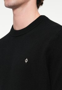 Fekete kötött pulóver kerek nyakkal, bal mellrészen egy kis fehér logóval. Az anyag textúrája lágy és sima.