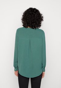 Blusa a maniche lunghe in tessuto verde, con orlo arrotondato, dettaglio plissettato sul retro e polsini con bottoni. Indossata con pantaloni neri.