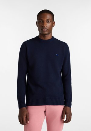 Navy plavi pleteni pulover s rebrastim izrezom i manžetama. Ima mali plavi logotip na prsima. Slim fit dizajn, uparen s ružičastim hlačama.