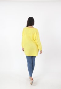 Übergroßer gelber Strickpullover mit langen Ärmeln und weitem Halsausschnitt, kombiniert mit blauen Skinny-Jeans und durchsichtigen Blockabsatzschuhen.
