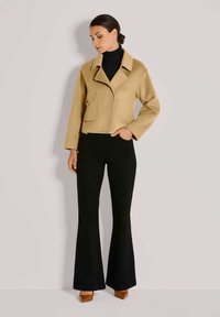 Frau mit einem hellbraunen, cropped Mantel über einem schwarzen Rollkragenpullover, schwarzen Schlaghosen und braunen spitzen Schuhen, die vor einem einfarbigen Hintergrund steht.