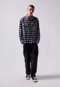 Jaunas vyras stovi, dėvintis juodai baltą languotą marškinį, juodas kelnes ir juodai baltus sportinius batelius prieš paprastą foną.