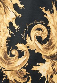 Χρυσά φύλλα ακάνθου και στροβιλίσματα σε μπαρόκ στιλ σε μαύρο ύφασμα με το κείμενο «Versace Jeans Couture» σε γραφικό χαρακτήρα.