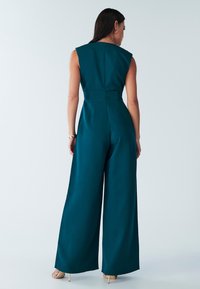 Combinaison sans manches en teal avec des jambes larges et une taille ajustée. Tissu lisse, semblable à du satin, avec une fermeture éclair discrète au dos et des coutures minimales.