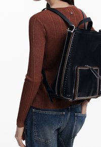 Osoba ubrana w brązowy sweter z prążkowanej dzianiny i niebieskie dżinsy, nosząca czarny skórzany plecak z plecionymi brzegami i przednią kieszonką na suwak.