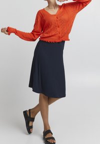 Punane kohev kardigan V-kaelusega, tekstuuriga muster ja volangiga äär, kombineeritud tumesinise ribakujulise midi-seeliku ja mustade avatud varvaste sandaalidega.