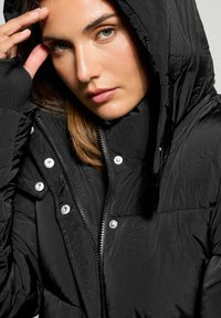 Svart puffjacka med huva, med dragkedja fram, tryckknappar, texturerad väv och elastiska ärmslut. Närbild av kragen.